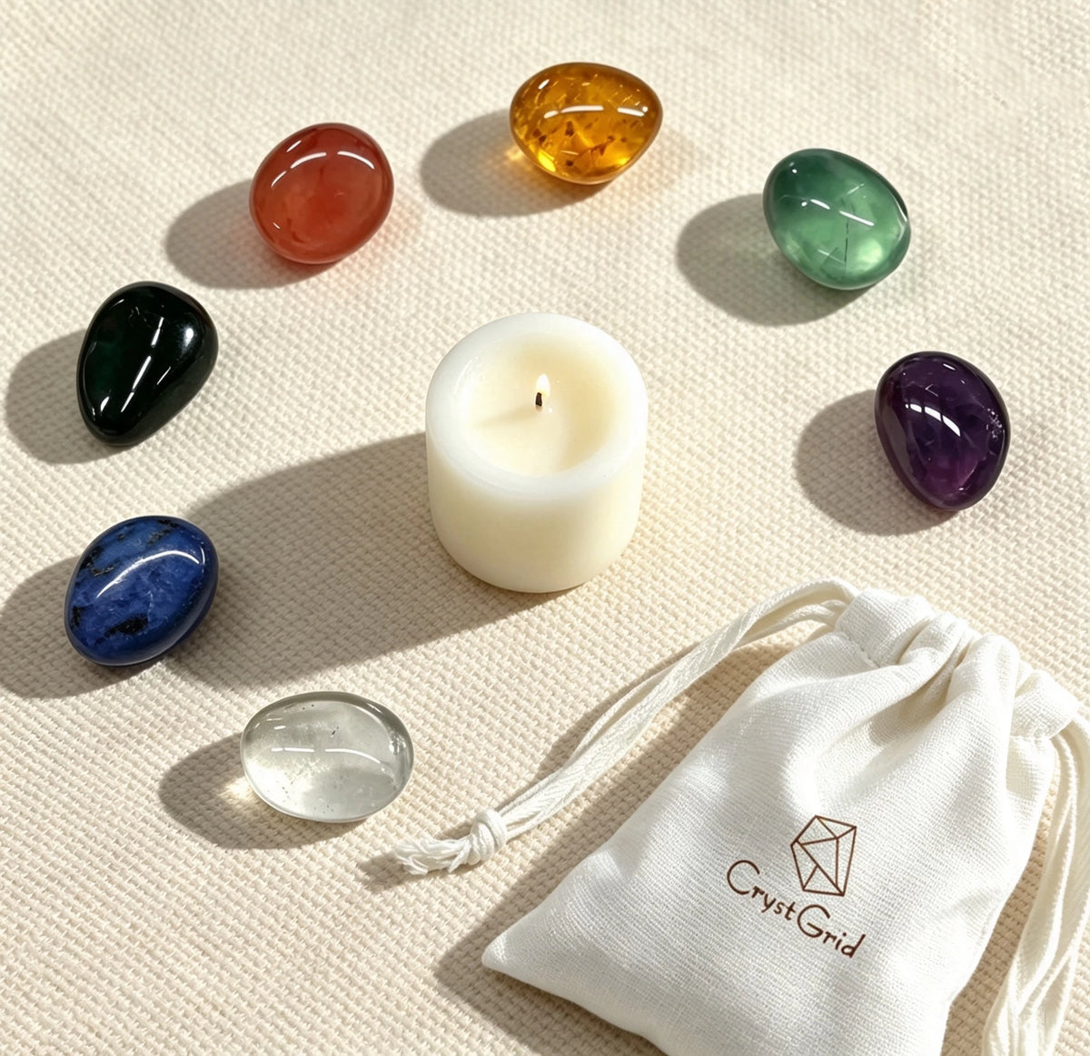 Seven chakra stones circled around a lit white candle on beige linen, CrystGrid pouch in the corner