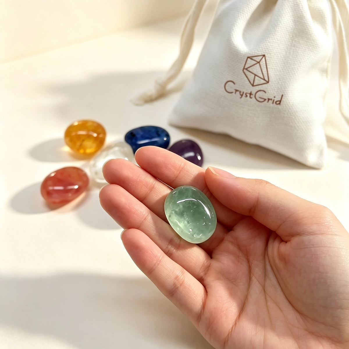 Hand holding one green chakra stone, other six stones and CrystGrid pouch blurred behind